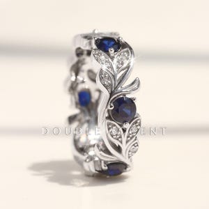May include: A silver ring with a leaf design, featuring round, dark blue gemstones and small clear stones. The ring's band has a repeating pattern of leaves and gemstones. The ring is displayed on a light background.