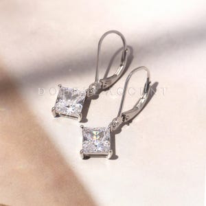 May include: A pair of silver-toned leverback earrings. Each earring features a square, clear, sparkling gemstone. The earrings are displayed on a light-colored surface. The text "DOUBLE ACCENT" is partially visible.