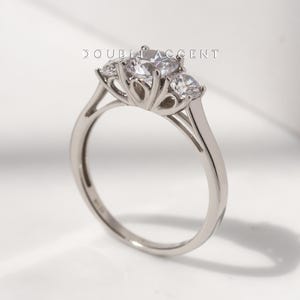 May include: A silver engagement ring with three clear, round-cut gemstones. The ring features a delicate band and a detailed setting that holds the stones. The text "DOUBLE ACCENT" is visible above the ring.