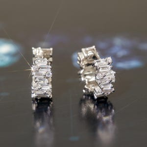 May include: A pair of silver-toned hoop earrings. Each earring features a series of rectangular and round-cut clear gemstones. The earrings are displayed against a dark, reflective background, with light reflecting off the gemstones.