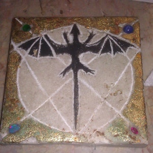 Puede incluir: Un azulejo cuadrado con una silueta negra de un dragón con las alas extendidas sobre un fondo blanco. El azulejo está decorado con pan de oro y tiene cuatro pequeñas piedras preciosas en las esquinas: dos verdes, una azul y una rosa.