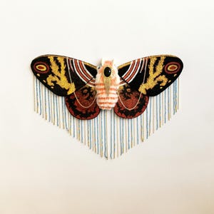May include: A decorative moth wall hanging featuring black, gold, and rust-colored wings with intricate patterns. The central body is white and orange with a black oval accent. Long, thin gold and light blue fringe hangs from the wings.