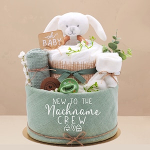Könnte beinhalten: Ein grüner und weißer Baby-Geschenk-Korb mit einem weißen Plüsch-Hasen, einem grünen Waschlappen, einem weißen Handtuch und einem Schild mit der Aufschrift "oh baby". Der Korb ist mit Grünpflanzen und einem grünen Band verziert. Der Text "New to the Nachname Crew" ist auf den Korb gedruckt.