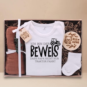 Könnte beinhalten: Ein weißer Baby-Strampler mit einem schwarzen Traktor-Motiv und dem Text "Ich bin der Beweis dass Papa nicht nur Traktor fährt". Der Strampler befindet sich in einer Geschenkbox mit einer braunen Musselin-Decke, einem Paar weißen Socken und einem Holzschild mit dem Text "Hello World".