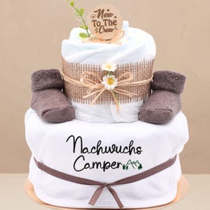 Könnte beinhalten: Eine zweistöckige Windeltorte, verziert mit einem braunen Juteband, weißen Gänseblümchen und braunen Babysöckchen. Die obere Etage hat ein Holzschild mit der Aufschrift "New To The Crew". Die untere Etage hat einen weißen Strampler mit dem Text "Nachwuchs Camper" und einer grünen Berggrafik.