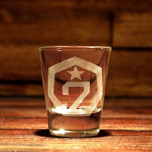 Puede incluir: Un vaso de chupito de vidrio transparente con un diseño grabado en blanco de una estrella y el número 7 dentro de un hexágono. El vaso de chupito está sobre una superficie de madera.