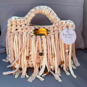 May include: A crocheted handbag in cream and dusty pink fabric yarn featuring a top handle, a gold-toned metal plaque labelled Boho Loops, and a round paper tag with the text boho loops. The bag is decorated with long fabric fringe and wooden beads.
