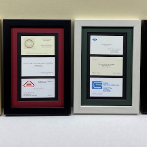 May include: Four framed displays of business cards, each with a different color frame. The frames are in a row from left to right: natural wood, black, white, and black. The business cards are all different, with some featuring logos and others featuring signatures.