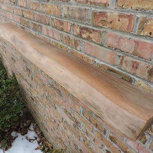 May include: A light brown, rectangular wooden shelf mounted on a brick wall. The shelf has a natural, unfinished look with visible wood grain. The brick wall is a mix of red, brown, and yellow bricks.