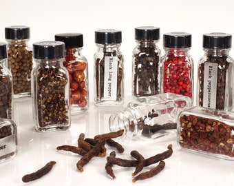 May include: A collection of small glass jars filled with various spices, including black peppercorns, pink peppercorns, long pepper, and Sichuan peppercorns. The jars have black lids and labels with the names of the spices. Some of the spices are scattered on the table.