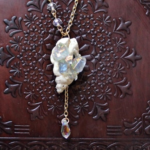 Op de afbeelding: Een gouden ketting met een hanger van witte en blauwe kristallen. De ketting hangt over een donkerbruin, reliëf ledergebonden boek. Amethist en andere kristallen zijn rond de ketting gerangschikt.