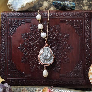 May include: A gold necklace with a white ammonite pendant and pearl accents hangs on a brown leather-bound journal with intricate embossed designs.