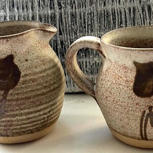 Könnte beinhalten: Zwei handgefertigte Keramikkrüge mit gesprenkelter beige-brauner Oberfläche. Jeder Krug hat ein dunkelbraunes Blumendesign und einen Henkel. Die Krüge eignen sich zum Servieren von Getränken.