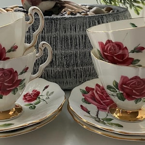 Op de afbeelding: Een set van zes vintage theekopjes en schotels. De kopjes en schotels zijn wit met een rood rozenbloemmotief en gouden randen. De theekopjes hebben een unieke handgreepvorm en staan op een gouden voet.