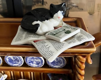 Może przedstawiać: Ceramiczna figurka czarnego i białego kota spoczywającego na talerzach w kształcie gazety. Talerze mają nadrukowany tekst i małą kartę z napisem "Rington's Dividend Card". Część dekoracyjnej półki.