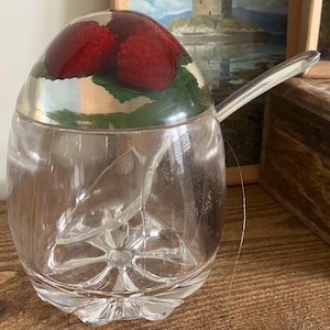 Könnte beinhalten: Eine transparente Zuckerdose aus Glas mit silbernem Rand und Löffel. Der Deckel hat eine Kuppel mit roten Himbeeren und grünen Blättern, die in klarem Harz eingeschlossen sind. Die Schale steht auf einer Holzoberfläche.