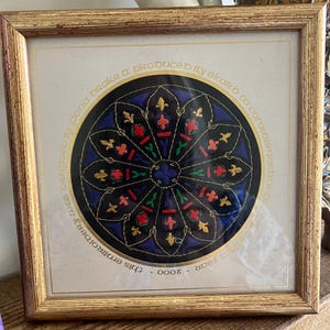 Puede incluir: Bordado enmarcado con un diseño circular sobre fondo negro y detalles coloridos. El diseño incluye elementos azules, rojos y dorados, enmarcado en un marco de madera dorada. Texto alrededor del círculo: "this embroidery..." y "2000".