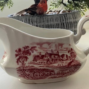 Könnte beinhalten: Eine weiße, gravierte Sauciere aus Keramik mit einem roten und weißen Blumendesign. Die Sauciere hat einen Griff und einen Ausguss. Das Design zeigt ein Häuschen mit rotem Dach und einen Baum.