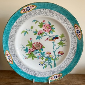 May include: A round, white ceramic plate with a teal border and floral design. The plate features a central image of a bird perched on a flowering branch with pink and red blossoms. The border has a repeating pattern and decorative accents.