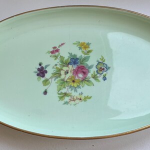 May include: An oval-shaped ceramic serving platter with a smooth, pale mint green glaze. The centre features a detailed floral bouquet illustration with pink, yellow, blue, and purple flowers and green leaves. The rim is finished with a thin gold band.