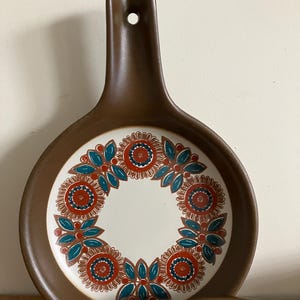 May include: Brown ceramic spoon rest with a handle and a circular design. The center of the spoon rest is white, and it features a floral pattern in red, teal, and brown. The handle has a hole for hanging.