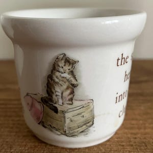 Könnte beinhalten: Ein weißer Keramik-Eierbecher mit abgerundeter Oberseite und einer detaillierten Illustration eines Kätzchens. Das Kätzchen sitzt auf einer Holzkiste. Der Text auf der Seite des Bechers lautet "the he into ch".