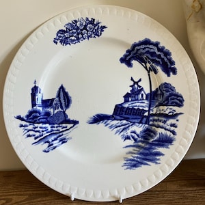 May include: White ceramic plate with a blue landscape design. The plate features a church, windmill, and floral accents. The rim has a textured pattern. A decorative piece for serving or display.