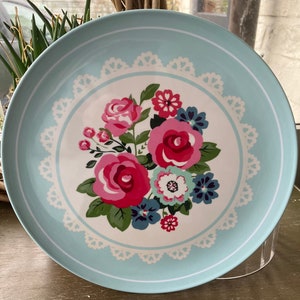 Könnte beinhalten: Ein runder, hellblauer Teller mit floralem Design. In der Mitte befindet sich ein Bouquet aus rosa und roten Rosen, blauen und weißen Blumen und grünen Blättern. Ein weißer, spitzenartiger Rand umgibt die Blumenanordnung.