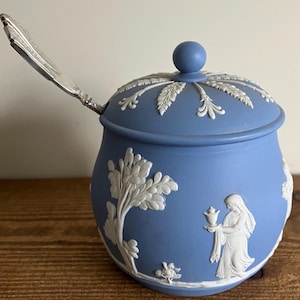 May include: A blue ceramic sugar bowl with a lid and a silver spoon. The bowl features white relief designs of trees, figures, and floral patterns. The lid has a round knob and a decorative edge.