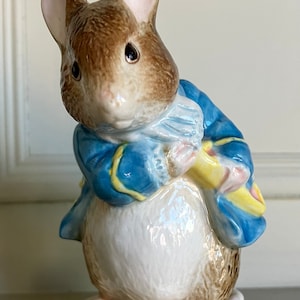 Puede incluir: Figura de cerámica de un ratón con una chaqueta azul con ribetes amarillos y una bufanda a juego. El ratón tiene pelaje marrón, orejas rosas y sostiene un objeto amarillo y rosa. La figura está sobre una base beige.