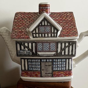 Op de afbeelding: Een keramische theepot in de vorm van een gebouw van twee verdiepingen met een rood pannendak en een schoorsteen. De buitenkant heeft zwart vakwerk, witte muren en blauw getinte ramen. Een bordje aan de voorzijde leest The Castle Inn, met een houten deur.