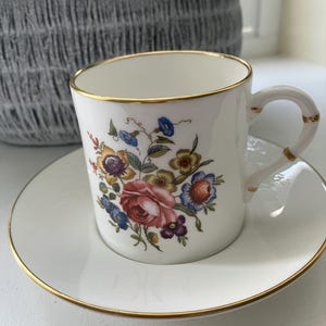 Puede incluir: Juego de taza y platillo de té blanco con ribete dorado. La taza de té presenta un diseño floral con flores rosas, azules, amarillas y moradas. El asa y el borde de la taza y el platillo tienen un ribete dorado.