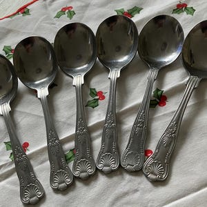 Op de afbeelding: Zes roestvrijstalen soeplepels op een wit tafelkleed met een hulst- en bessenpatroon. De lepels hebben een afgeronde kom en een sierlijk handvat.