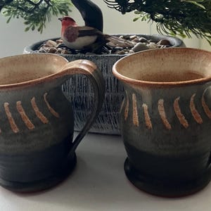 May include: Two handmade ceramic mugs with a dark grey and brown glaze. Each mug features a curved handle and a series of vertical, light brown stripes. The interior of the mugs is a light brown colour. The mugs are sitting on a white surface.