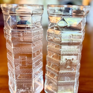 May include: Two clear glass vases with a geometric design. The vases have a rectangular shape with textured, patterned details. The design includes diamond shapes and rectangular patterns. The vases are displayed on a wooden surface.