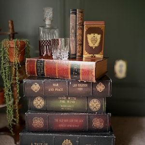 May include: A stack of antique books with titles like "War and Peace" and "Gone with the Wind." A glass decanter, a tumbler, and a decorative tin sit atop the books. The books are in various colors, including red, brown, and black.