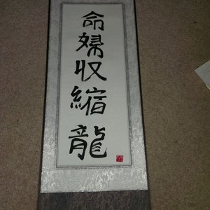 May include: A black and white scroll with Chinese calligraphy that reads "合気道" which translates to "Aikido". The scroll is mounted on a brown wooden dowel and has a silver background.