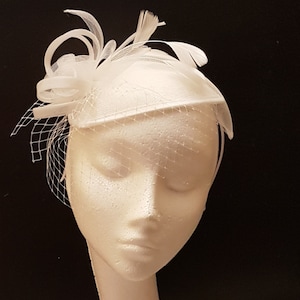Könnte beinhalten: Ein weißer Fascinator mit Schleier, Federn und dekorativen Akzenten. Der Kopfschmuck wird auf einem Schaufensterpuppenkopf vor schwarzem Hintergrund präsentiert. Dieses elegante Accessoire eignet sich für Hochzeiten oder besondere Anlässe.
