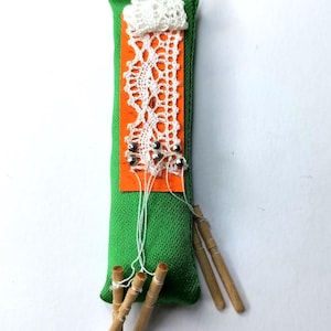 May include: A handmade green fabric ornament with a red ribbon loop. It features an orange panel with white lace and silver beads, and small wooden dowels attached with string. The ornament is approximately 5 inches long.