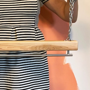 May include: A wooden swing with a stainless steel bar, suspended by metal chains. The swing is made of light-coloured wood and has a simple, modern design. The bar is approximately 61 cm long. The background is a wall with peach-coloured paint.