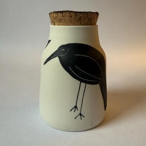 May include: A cylindrical off-white ceramic jar featuring a black graphic illustration of a bird with long legs and a pointed beak. The jar is topped with a natural brown cork stopper that fits securely into the opening at the top of the vessel.