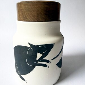 May include: A cylindrical ceramic jar with a matte white finish and a dark brown wooden lid. The exterior features a black graphic illustration of a small animal with pointed ears and a long tail, depicted in a dynamic, leaping pose against the white.