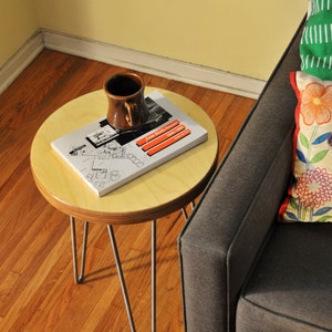 Puede incluir: Una peque&ntilde;a mesa auxiliar redonda de madera con patas de horquilla. La mesa tiene un libro y una taza de cer&aacute;mica marr&oacute;n en la parte superior. La mesa est&aacute; junto a un sof&aacute; gris con un coj&iacute;n de estampado verde y floral.