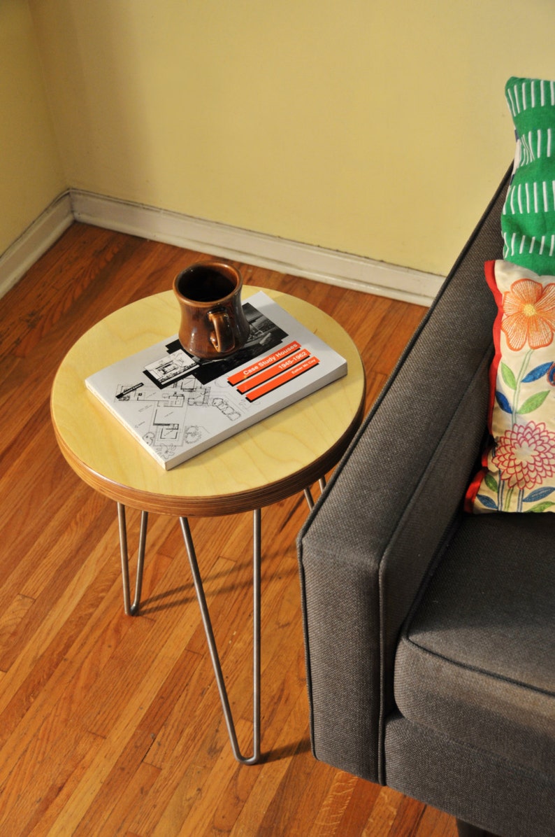 Puede incluir: Una peque&ntilde;a mesa auxiliar redonda de madera con patas de horquilla. La mesa tiene un libro y una taza de cer&aacute;mica marr&oacute;n en la parte superior. La mesa est&aacute; junto a un sof&aacute; gris con un coj&iacute;n de estampado verde y floral.