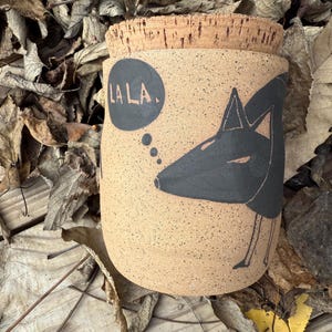 May include: A tan ceramic jar with a cork lid. The jar features a black illustration of a fox with the text "LA LA." in a speech bubble. The jar is sitting on a bed of dried leaves.
