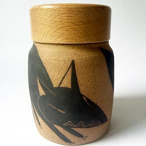 May include: A cylindrical ceramic jar with a speckled tan finish and a light-colored wooden lid. The jar features a black graphic illustration of a stylized animal with pointed ears and a jagged mouth design etched into the surface of the clay.