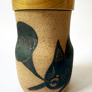 May include: A speckled tan ceramic canister with a smooth wooden lid. The canister features a black graphic of a cat with an open mouth and a speech bubble containing the text RAWR! The cat has pointed ears, whiskers, and visible claws.