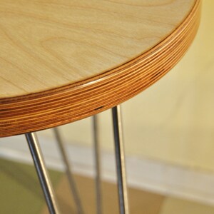 Puede incluir: Una mesa redonda de madera con acabado de madera natural y borde de madera contrachapada. La mesa est&aacute; sostenida por tres patas de horquilla de metal.