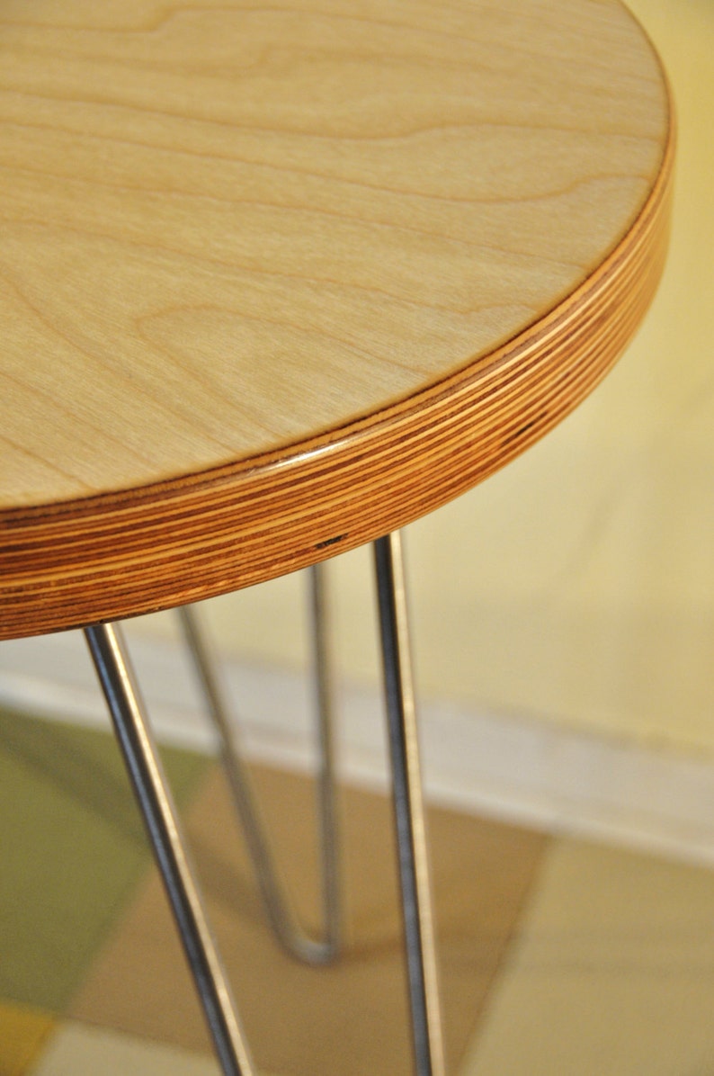 Puede incluir: Una mesa redonda de madera con acabado de madera natural y borde de madera contrachapada. La mesa est&aacute; sostenida por tres patas de horquilla de metal.