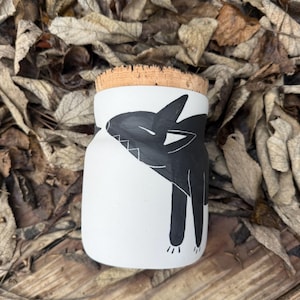 May include: A white ceramic jar with a cork stopper, featuring a black illustration of a stylized animal. The animal has pointed ears and a long tail. The jar is sitting on a wooden surface.
